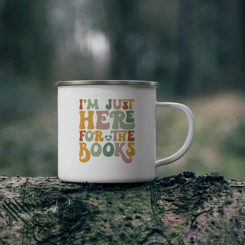A white metal camping mug on a log with the phrase "I'm just here for the books" in a vintage colorful font