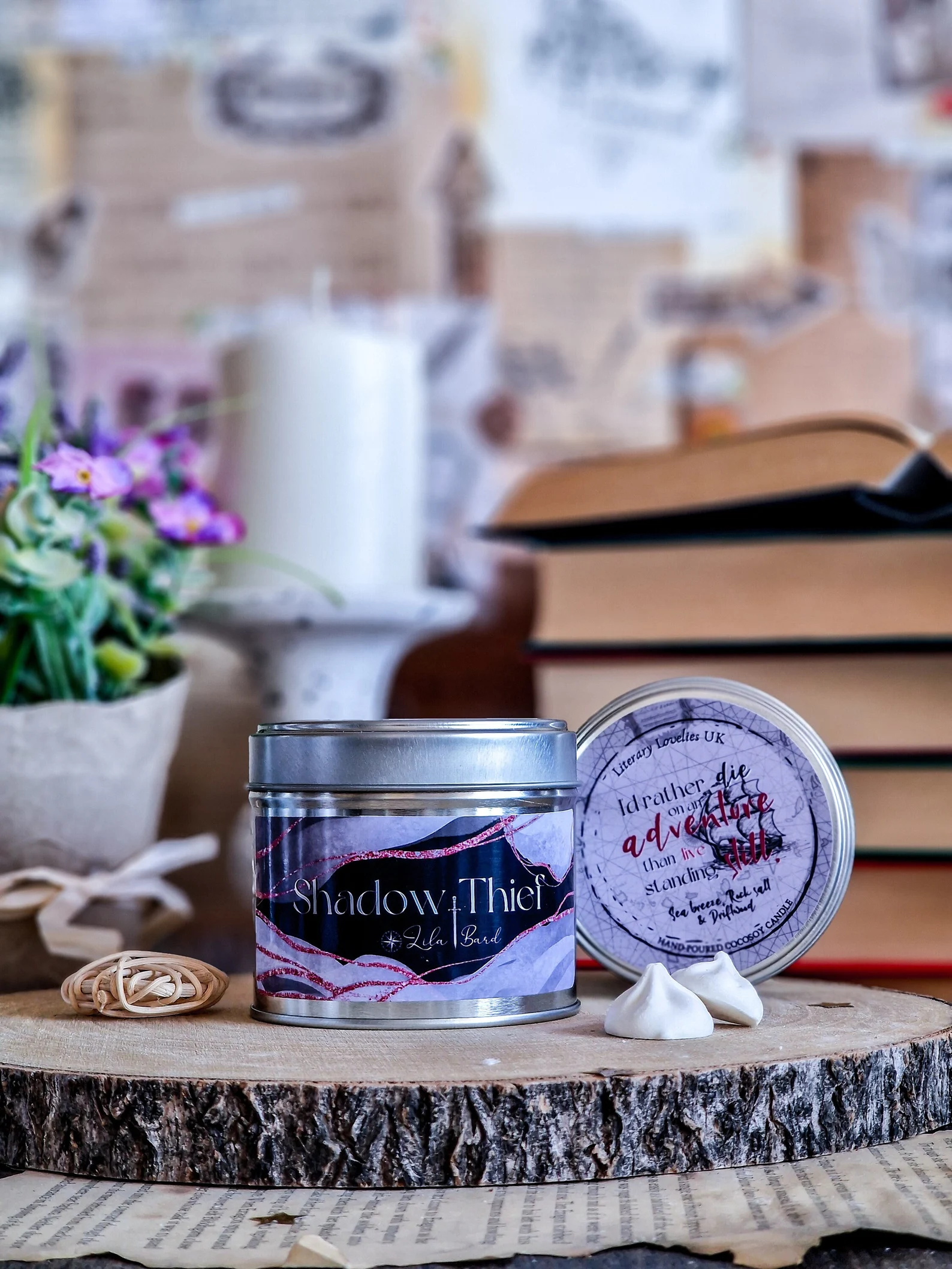 Two candles in tins with the label shadow thief + Lila Bard in front of flowers, a candle, and a stack of books.