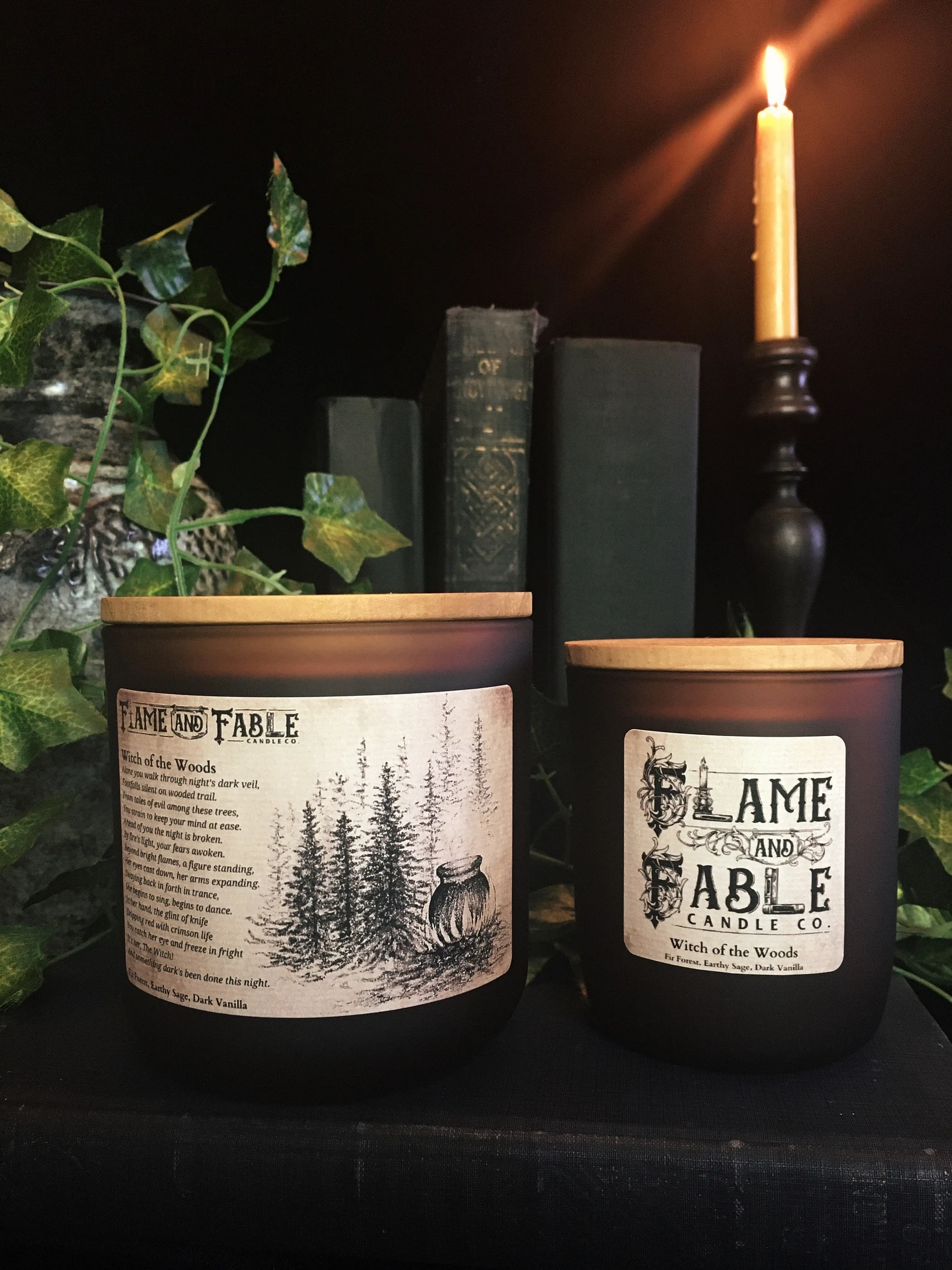 Two candles with the label Flame and Fable witch of the woods one large and the other small in front of a jar with a plant, three green books, and a candle on a candlestick.