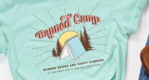 a photo of a teal t-shirt that says "Banned Camp: Banned Books and happy campers, if you don't like it, you can take a hike" and a graphic of a book standing spine-up like a tent surrounded by trees