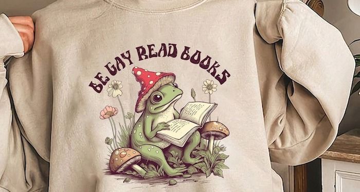 a photo of a sweatshirt with an illustration of a frog with a wizard hat reading a book and the text Be Gay Read Books