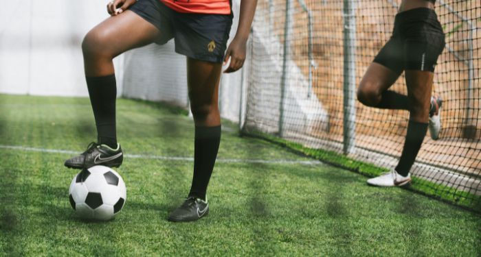 legs of two Black women on a soccer pitch, one with a leg perched on top of a soccer ball