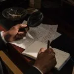 brown-skinned hands holding magnifying glass and looking at papers on a desk
