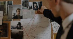 a man pinning a photo to a cork board with other images, evidence, and red string
