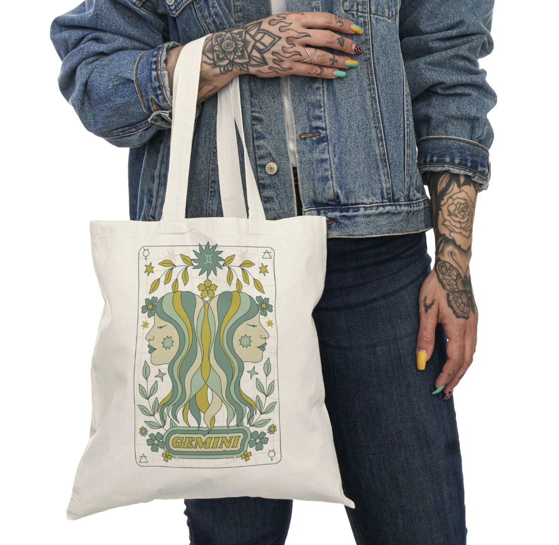 The torso of a tattooed person in a denim jacked holding an off-white canvas tote bag with a green and blue 1970s style illustration of two twin women and the word "Gemini"