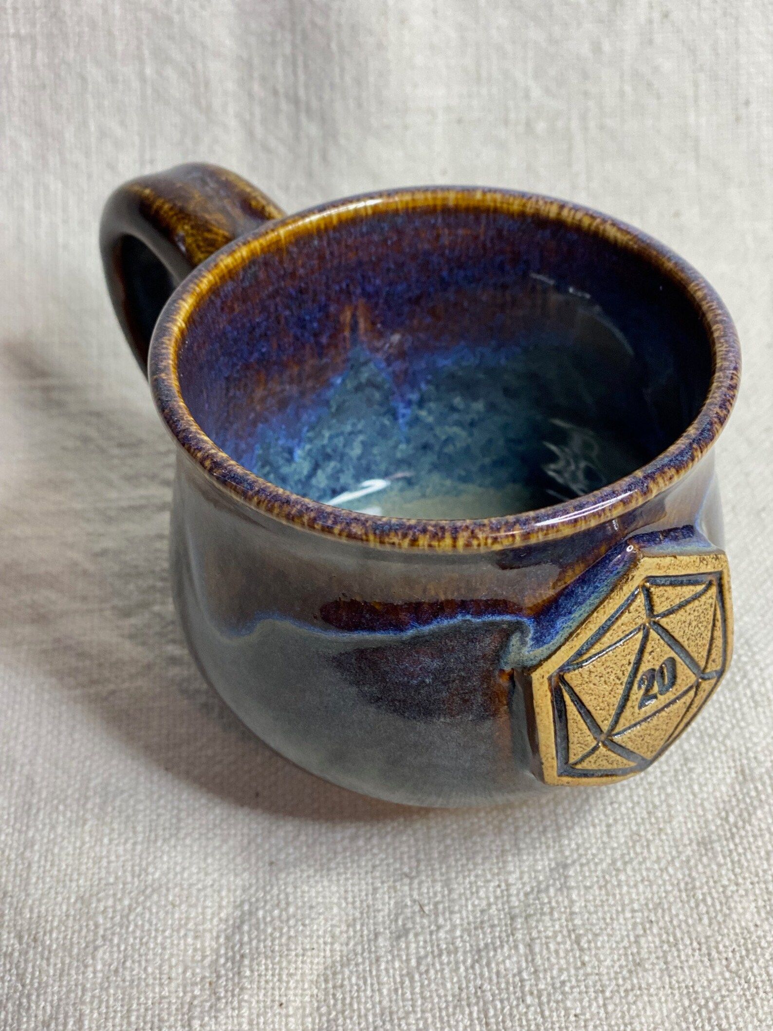A blue and purple ceramic mug with a natural 20 on a 20 sided dice on the front sits on white fabric.