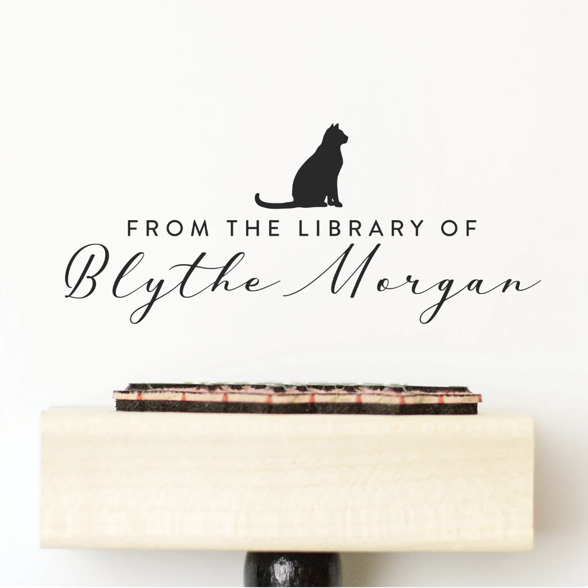 A rubber stamp on a wooden block below a stamp in black ink on a white page with an outline of a cat in profile and the text "From the library of Blythe Morgan"