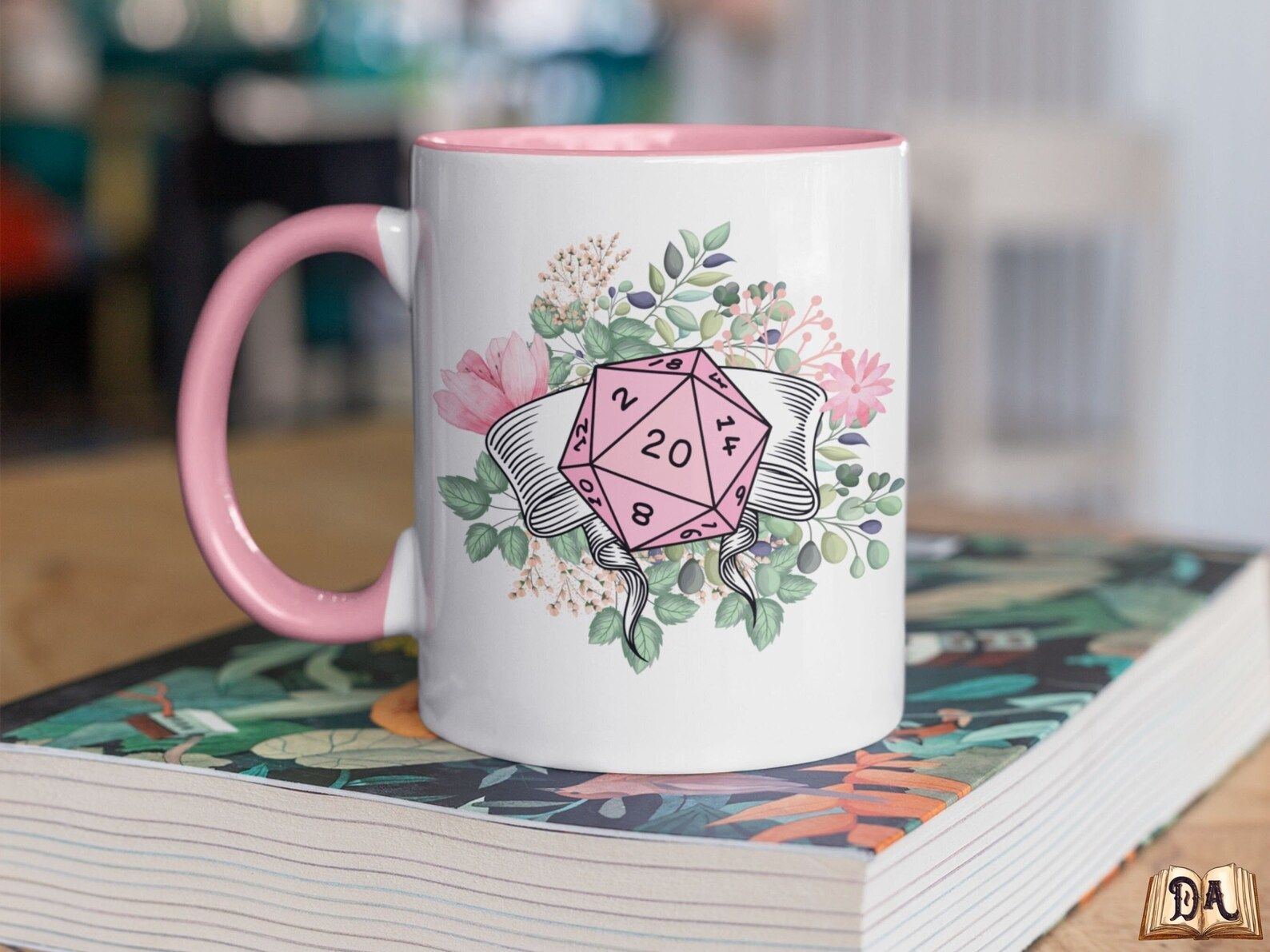 A white ceramic mug with a pink handle and interior has a 20 sided dice in front of flowers on the side. It sits on a book and a wooden table.