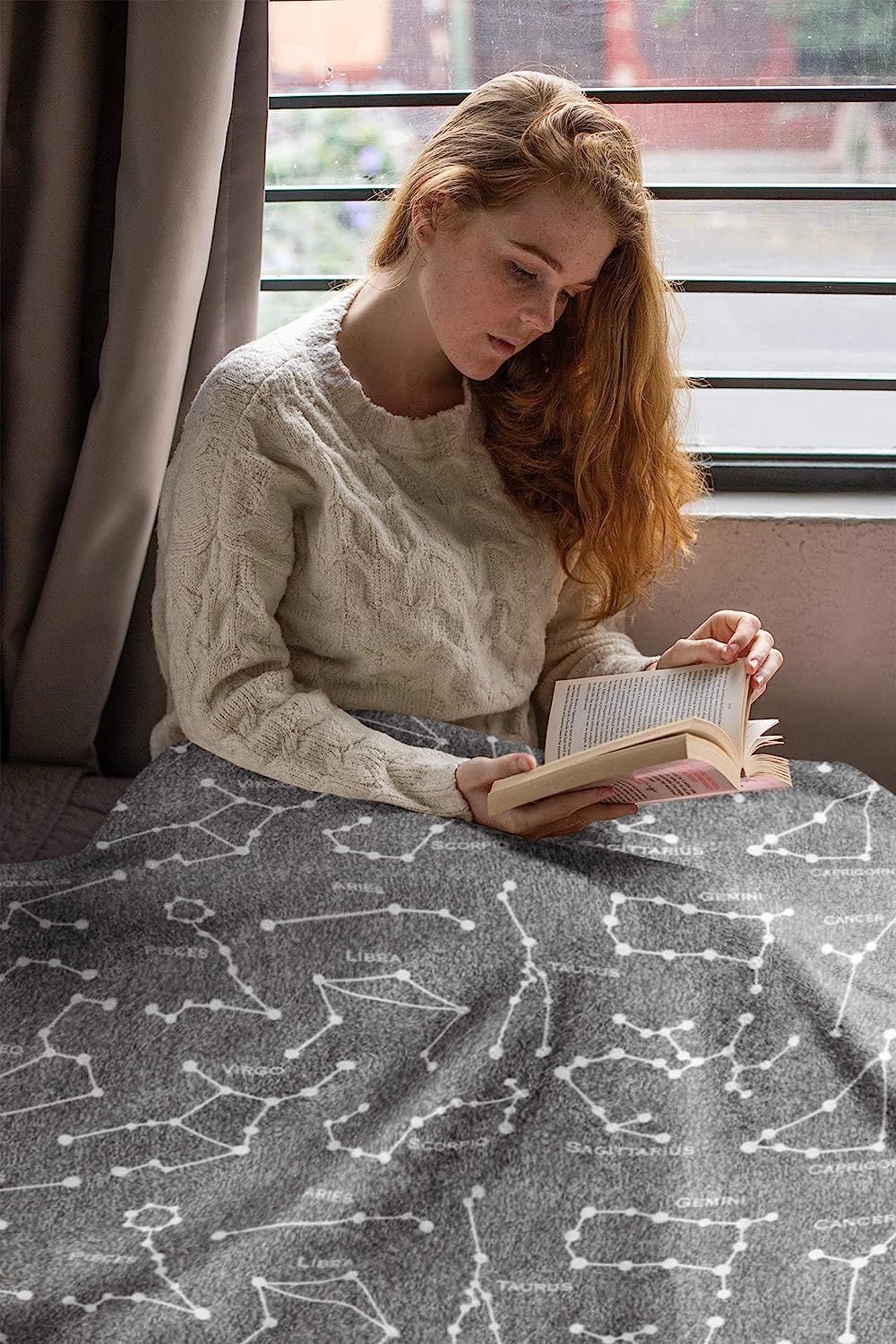 A person is reading while seated with a gray blanket covered in constellations spread across their lower half