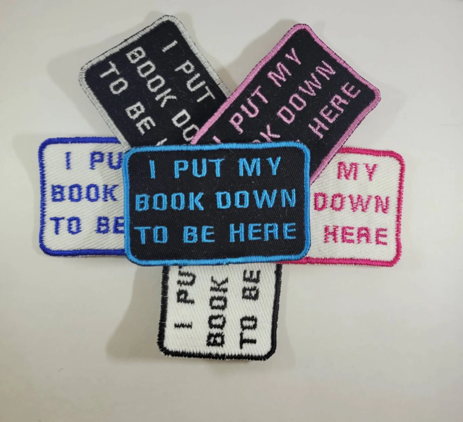 A pile of several small rectangular patches, in different color stitching, with the slogan, "I put my book down to be here."