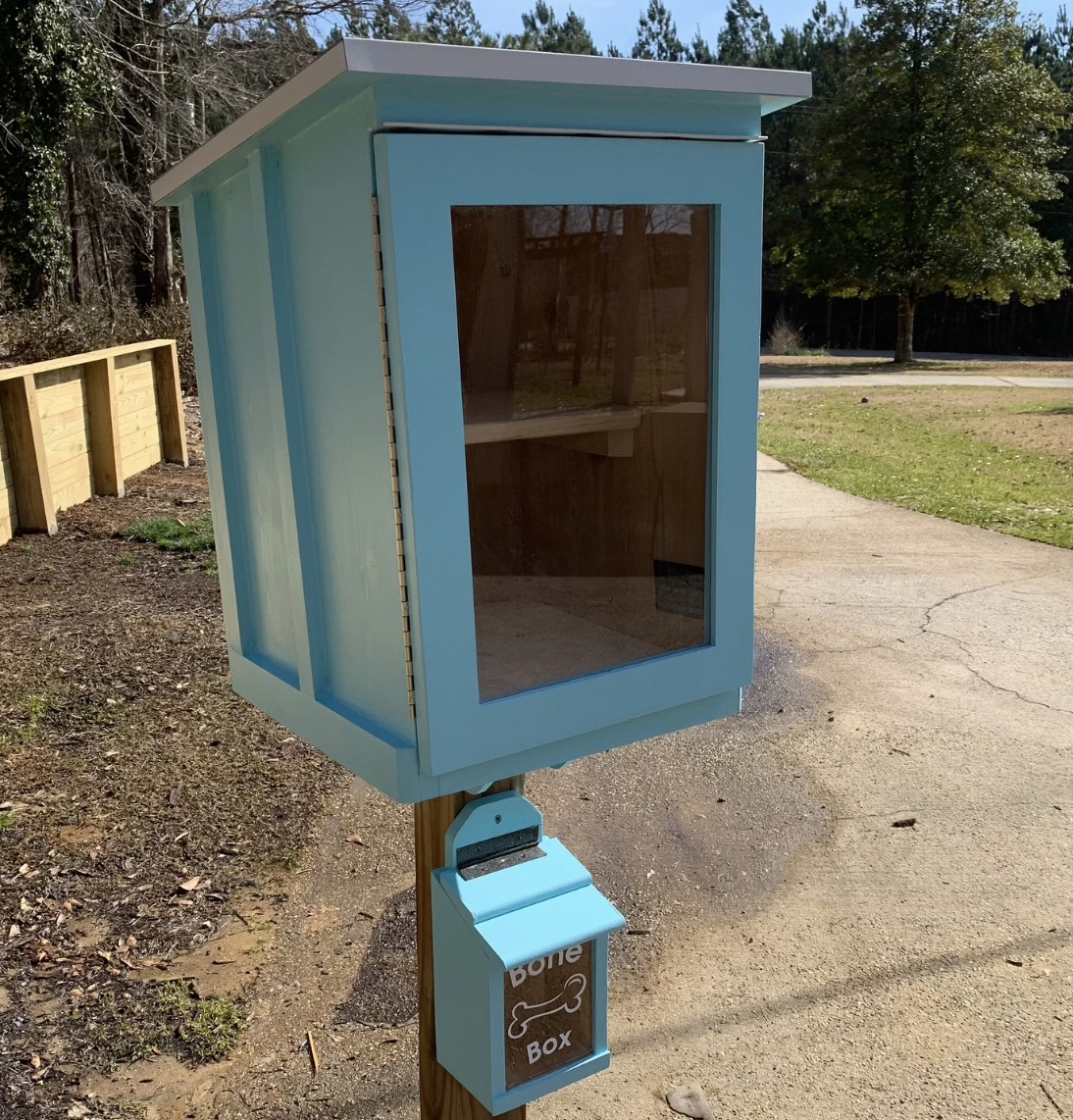 A wooden Little Free Library painted teal with a matching little wooden box underneath that says "Bone box"