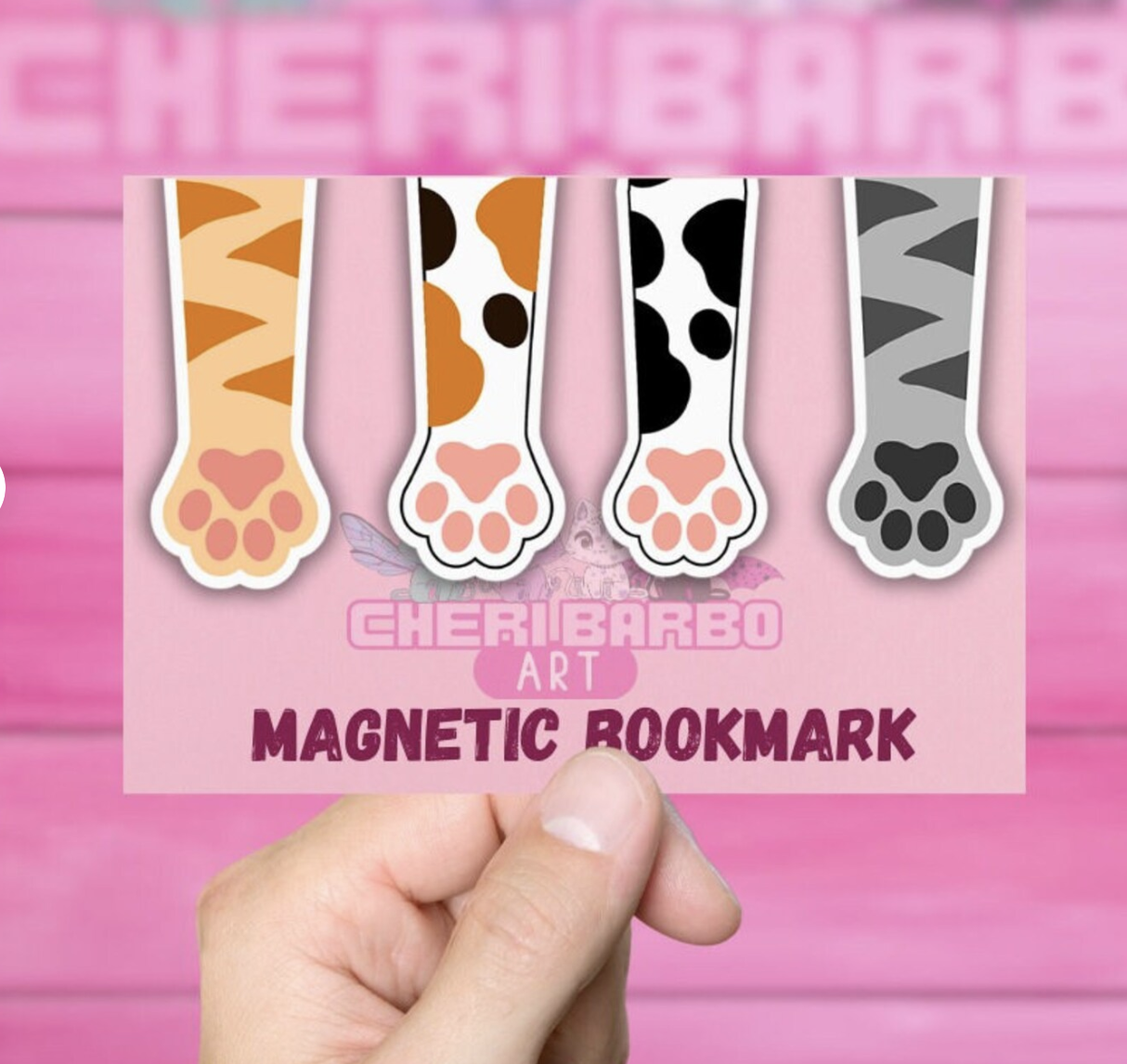 A hand holds a pink cardboard sheet with four magnetic cat paw bookmark clipped over the top. The paws are orange striped, calico, white with black spots, and gray striped. 