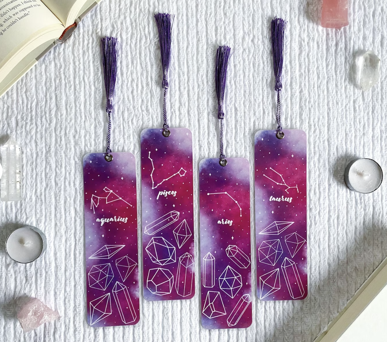 Four bookmarks with purple tassels, purple and pink watercolor designs and various zodiac signs and matching constellations on a white backdrop