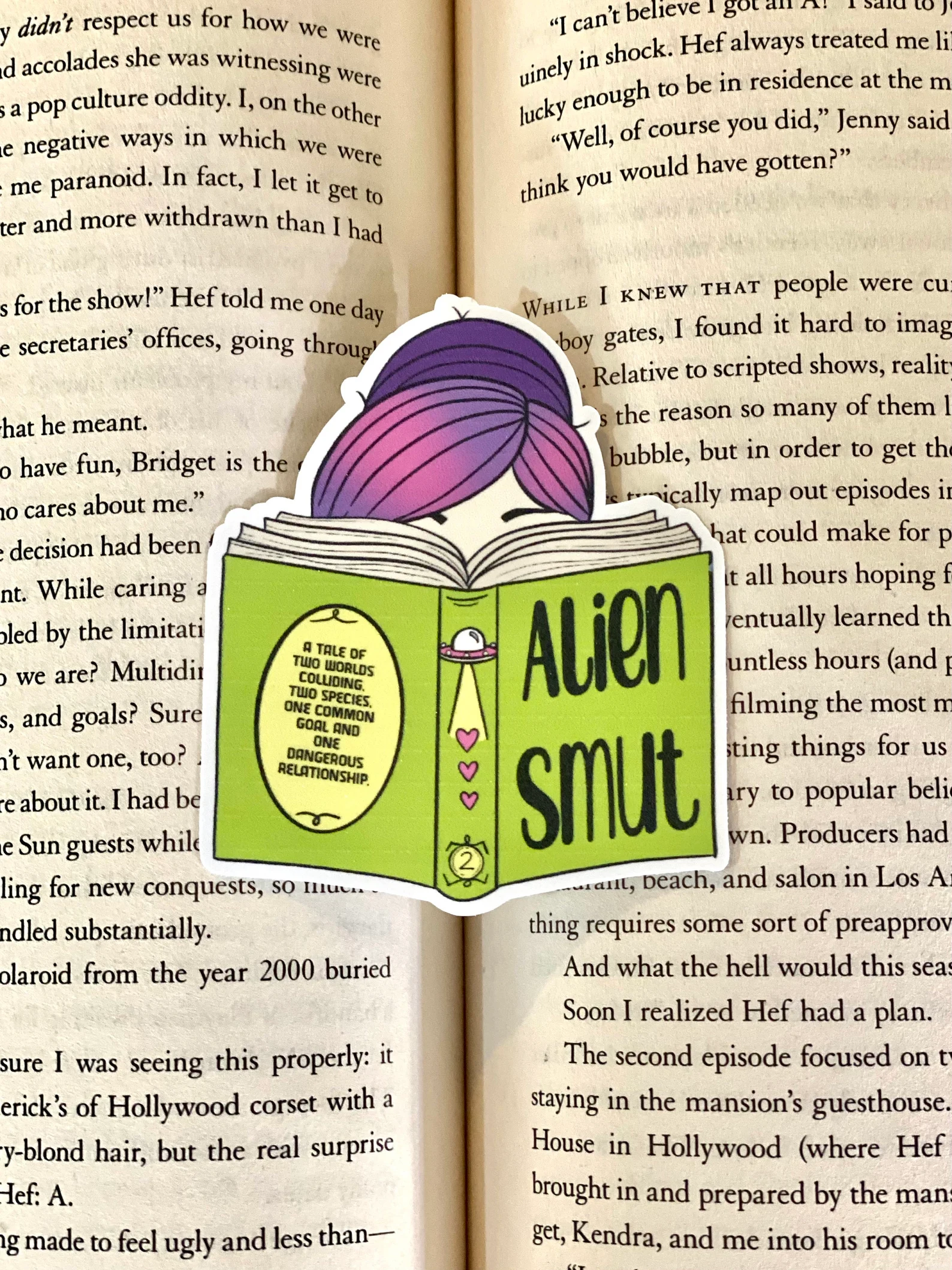 Image of a sticker featuring a person's head with purple hair and a neon green book that is titled "alien smut."