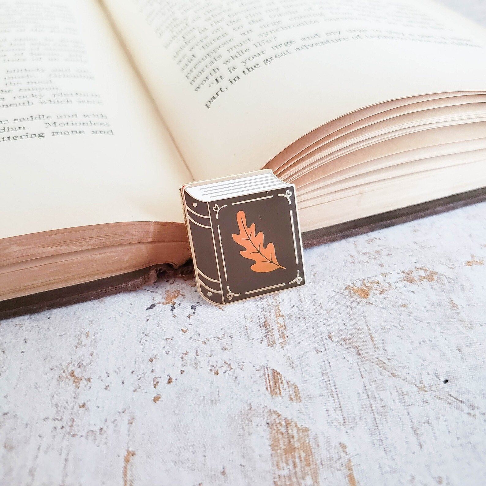 enamel pin of a book with an oak leaf on it