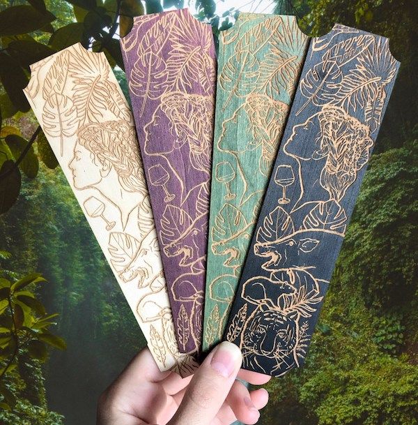 cream, purple, green, and blue wooden bookmarks with carved illustrations of a woman in profile plus an assortment of animals and plants