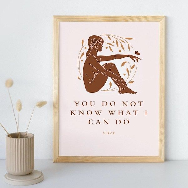 pale pink print in a wooden frame with a solid brown image of a woman seated with her knees drawn to her chest and the words "You Do Not Know What I Can Do"