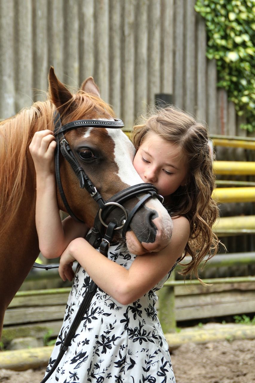 A young white girl with blonde hair, wearing a white dress with a black floral pattern, is hugging a chestnut pony with a white stripe down the front of its face