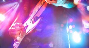 person with tattooed arms playing al electric guitar bathed in neon pink and blue light