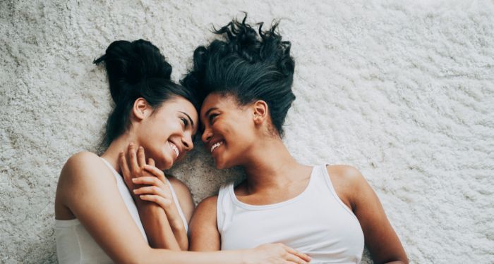 two brown-skinned women smiling while caressing each other in bed