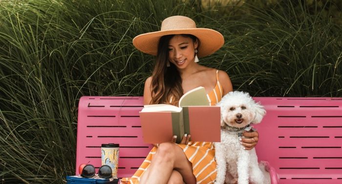 tan skinned woman of color sitting on a pink bench with a white dog; shes smiling and reading