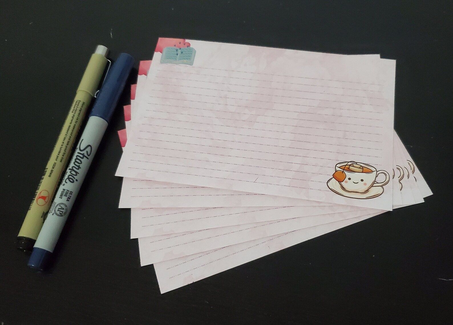 A small stack of lined notecards with an open book in one corner and a smiling tea cup in the other.