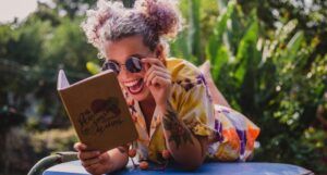 fair skinned woman with pom pom hairstyle laughing while reading a book and lying on top of a car