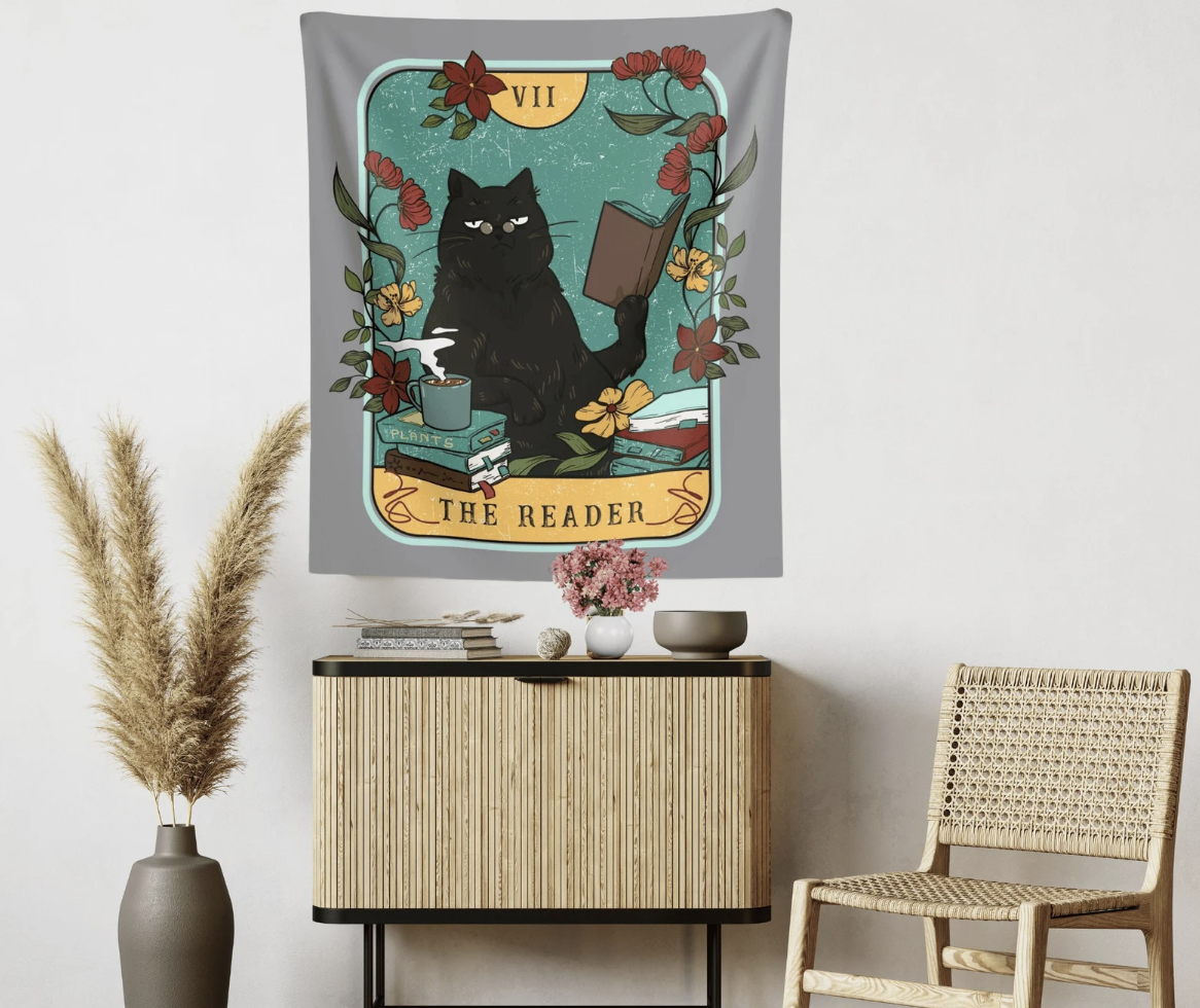 A fabric hanging tapestry designed to look like a tarot card that says "The Reader" with an illustration of a black cat reading a book