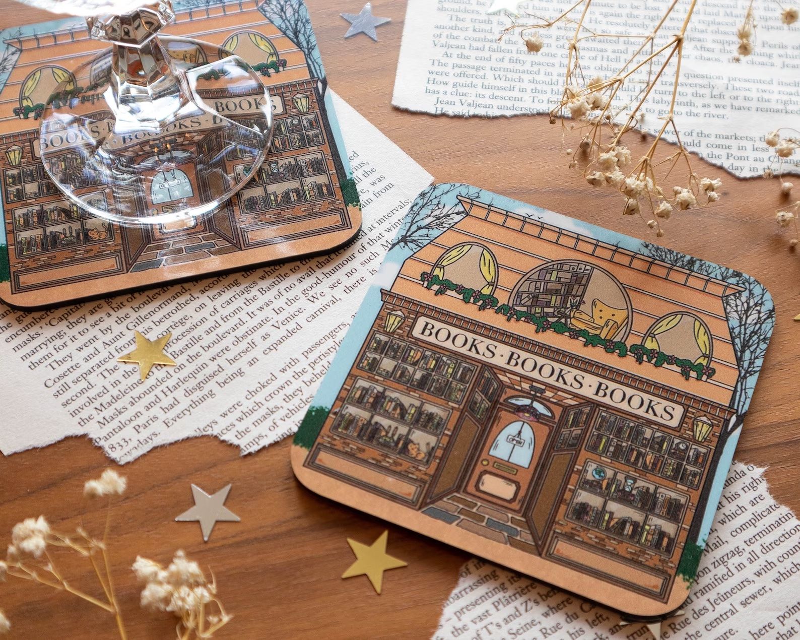 Coaster with a bookstore on it is on top of a table with ripped book pages, stars, and flowers.