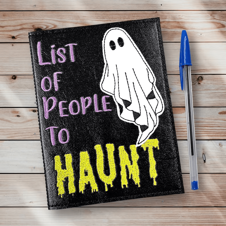 Photo of a black notebook with a pen placed next to it. The notebook has a sheet-ghost in the cover and the text, in purple and yellow, List of people to haunt. 