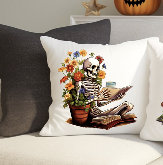 A white throw pillow with an image of a skeleton seated and reading a book next to a big pot of wildflowers