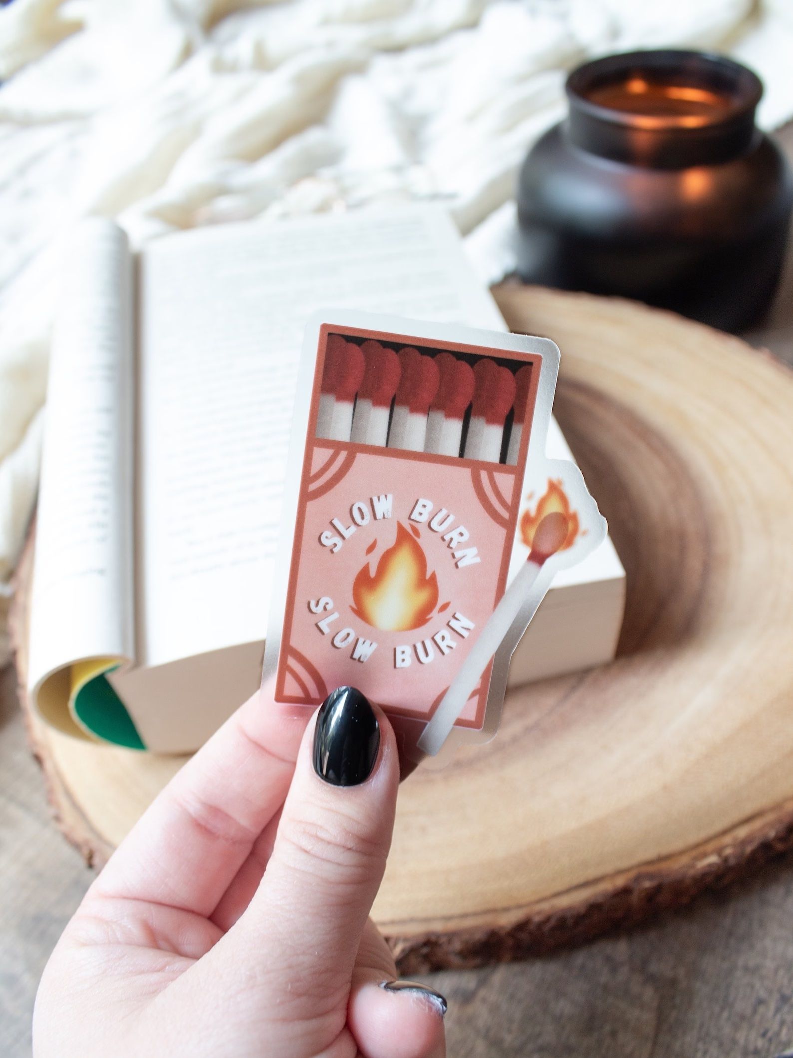 Slow Burn Book Trope Match Box Sticker held in front of a book on a wooden cutting board and wooden table. A candle and blanket sit in the background.