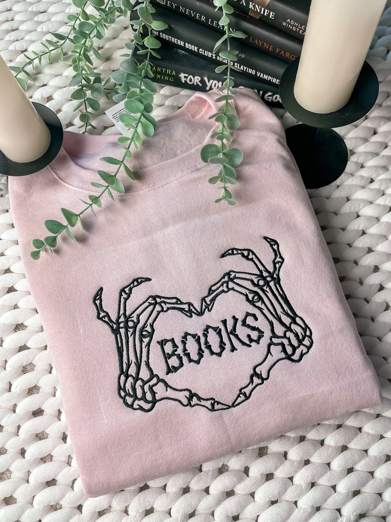 Image of a lilac colored sweatshirt. Embroidered on it are two skeleton hands in the shape of a heart with the word "books" between them. 