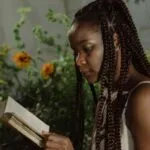dark brown-skinned woman with braids reads a boot in a lush green garden