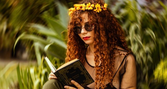 fair skinned white woman with long, curly red hair and a crown of yellow flowers. She's reading a book and the background is green with a swirling effect