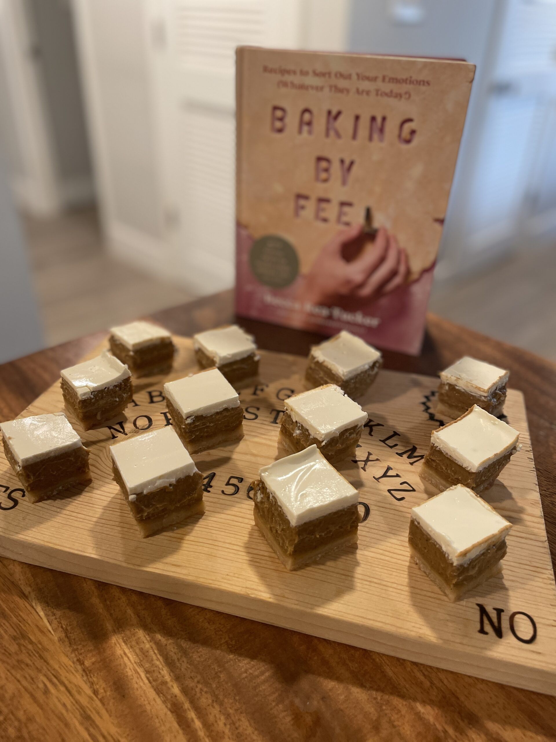 Twelve layered pumpkin pie bars with sour cream topping on a wooden cutting board designed to look like a ouija board, sitting on a wooden table next to the cookbook Baking By Feel