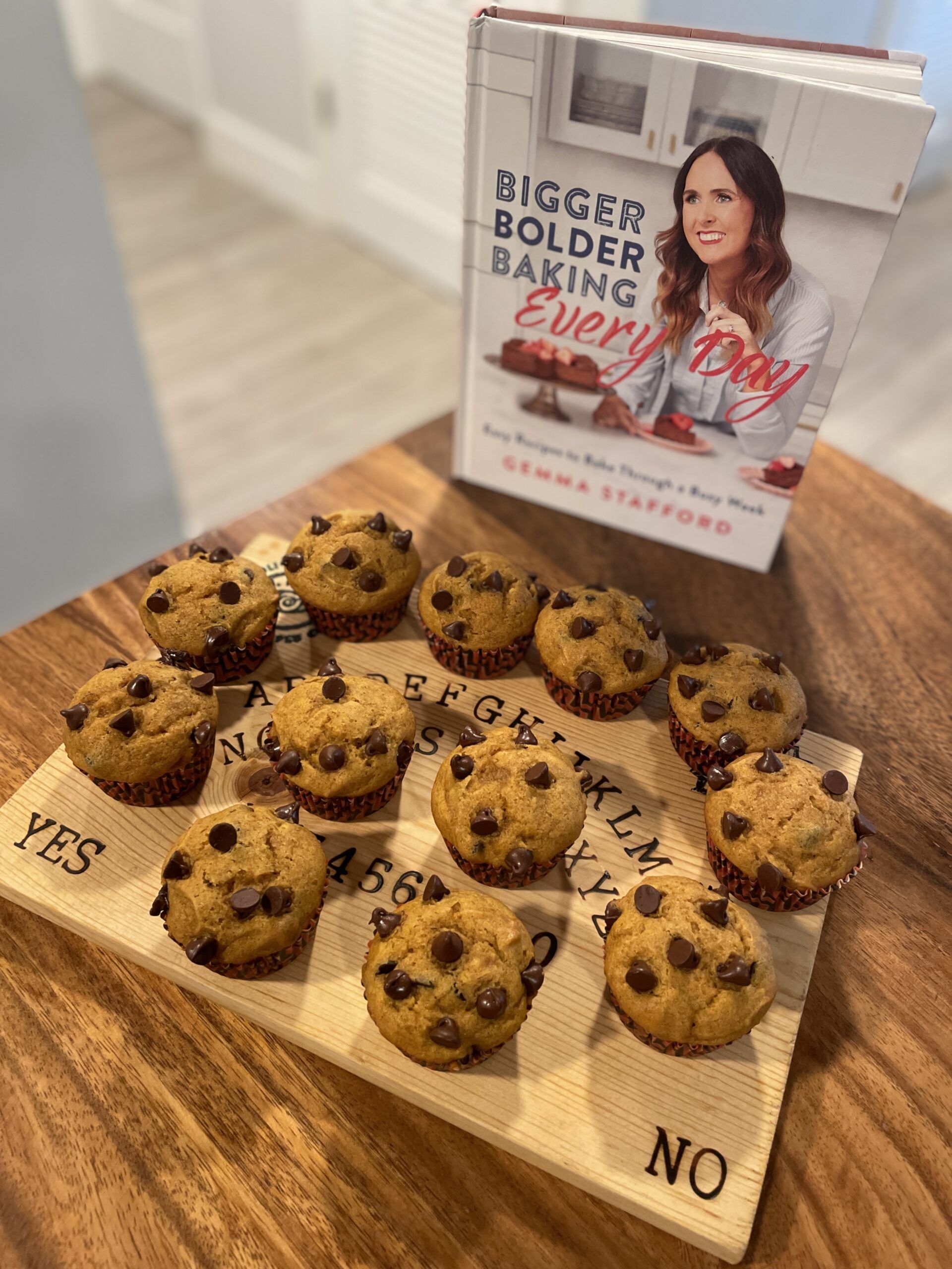 A dozen pumpkin muffins with chocolate chips on top displayed on a wooden cutting board that looks like a ouija board sitting on a wooden table by the cookbook Bigger Bolder Baking Every Day