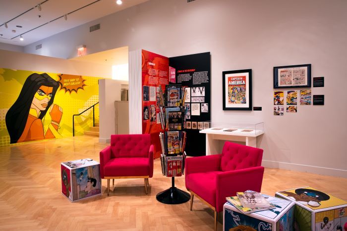 Photo of JewCe exhibit, showing two armchairs, a rack of comics, and comic covers on display