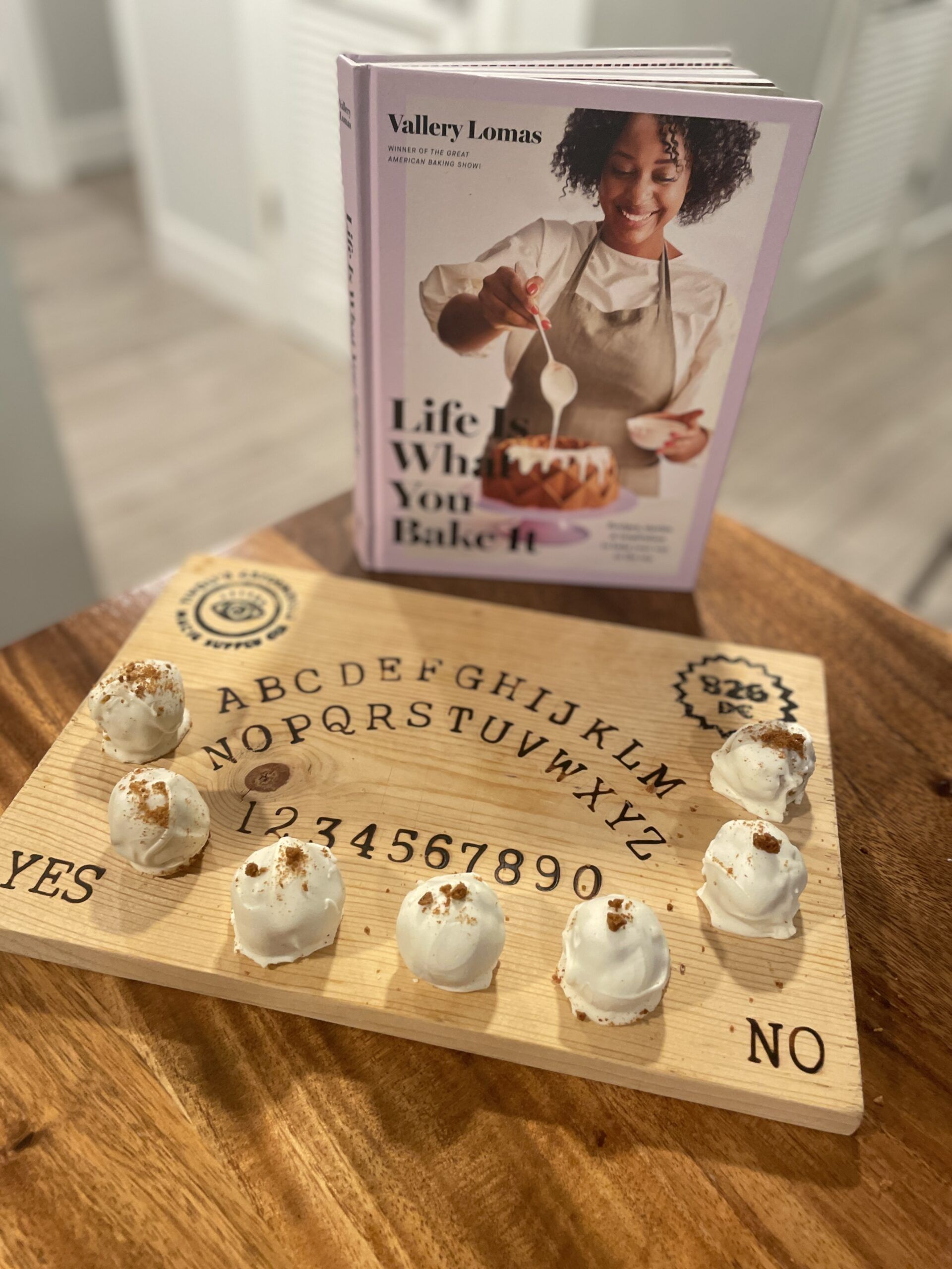 Seven white chocolate covered truffles sprinkled with gingersnap crumbs on a wooden ouija cutting board on a wooden table next to the cookbook Life is What You Bake It