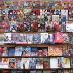 A person with short dark hair, wearing a red coat, stands in front of a wall displaying many different comic books