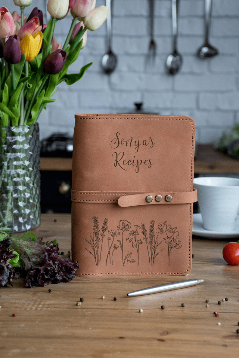 Photo of a leather notebook with a leather strap that goes around it and closes with three claps, with some flowers and text engraved on the cover