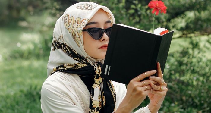 light tan skinned wman with head scarf reading a book with a red flower sticking out of it like a bookmark