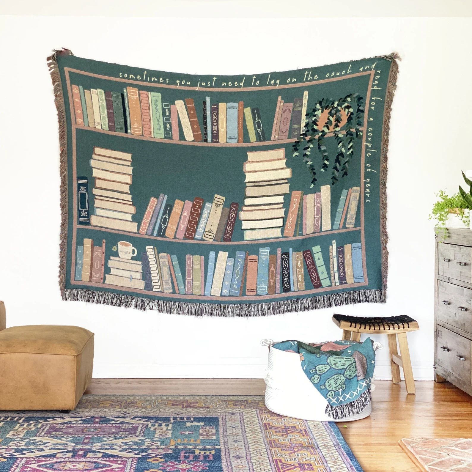 Book Lover Throw Blanket hung on a white wall in front of a couch, basket, and side table.