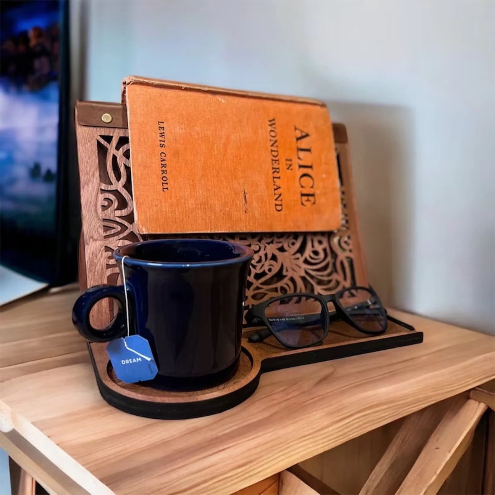 Wooden book tray with a mug, book, and glasses on it sits on top of a wooden table. 