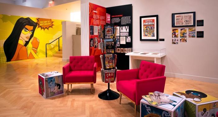 Photo of JewCe exhibit, showing two armchairs, a rack of comics, and comic covers on display
