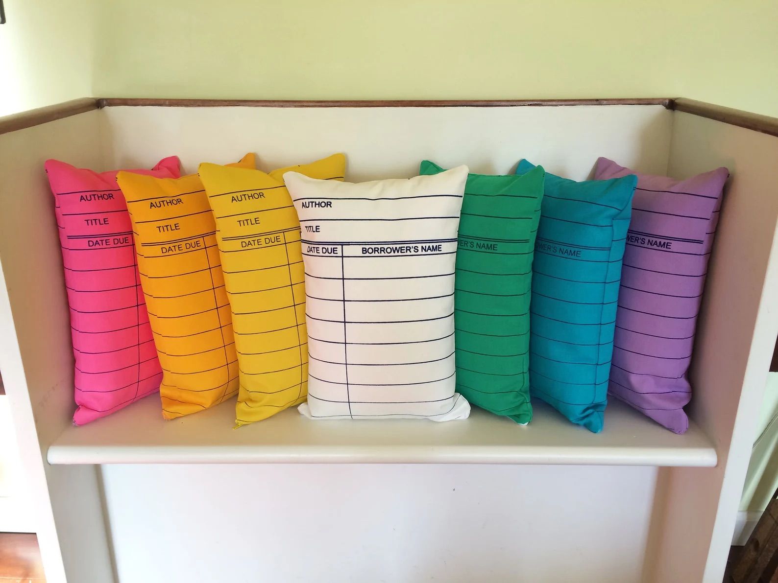 Seven Library Card Pillows in a rainbow of colors are on a white bench.