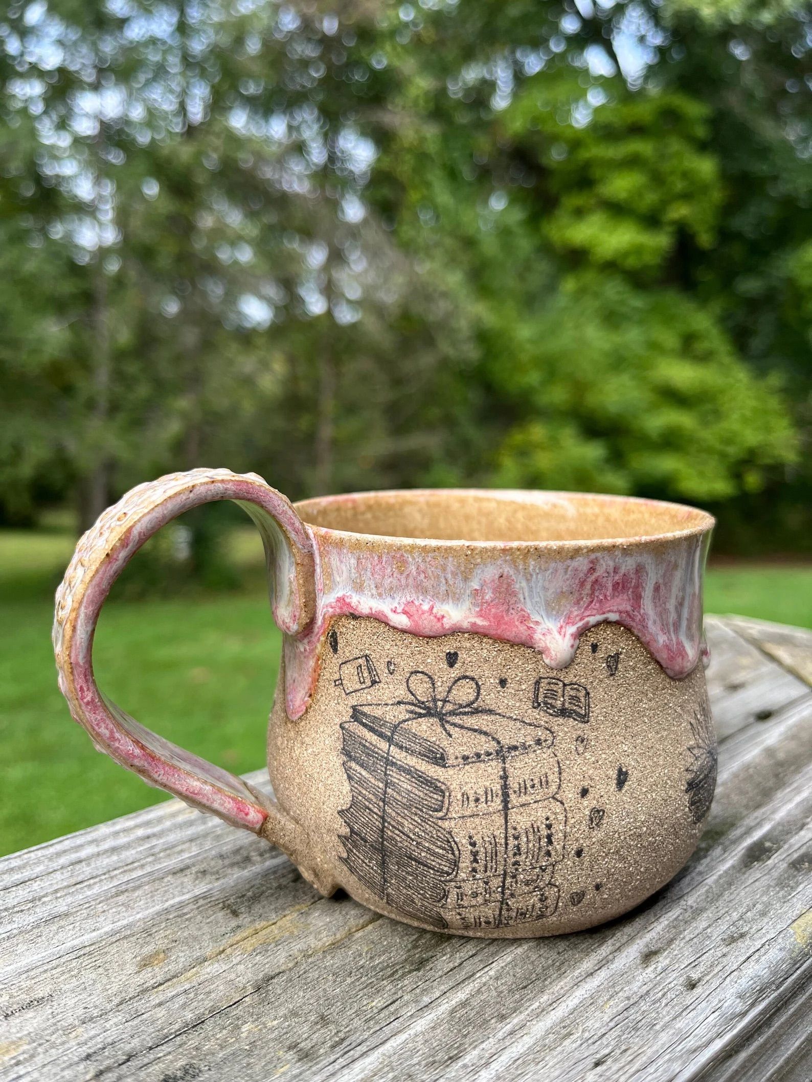 A ceramic mug with a stack of books on the side is on a table outside in the woods. 