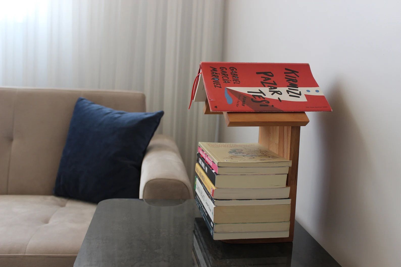 Wooden book home with a stack of books in it and an open book on top is on a table in front of a couch. 