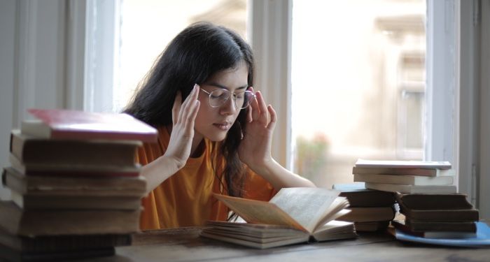 a lighter-skinned woman of color with dark hair has her hands to her temples while she sits on a desk full of books