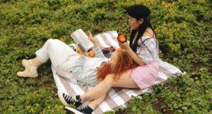 an Asian and white woman sitting and reading a book on a picnic blanket